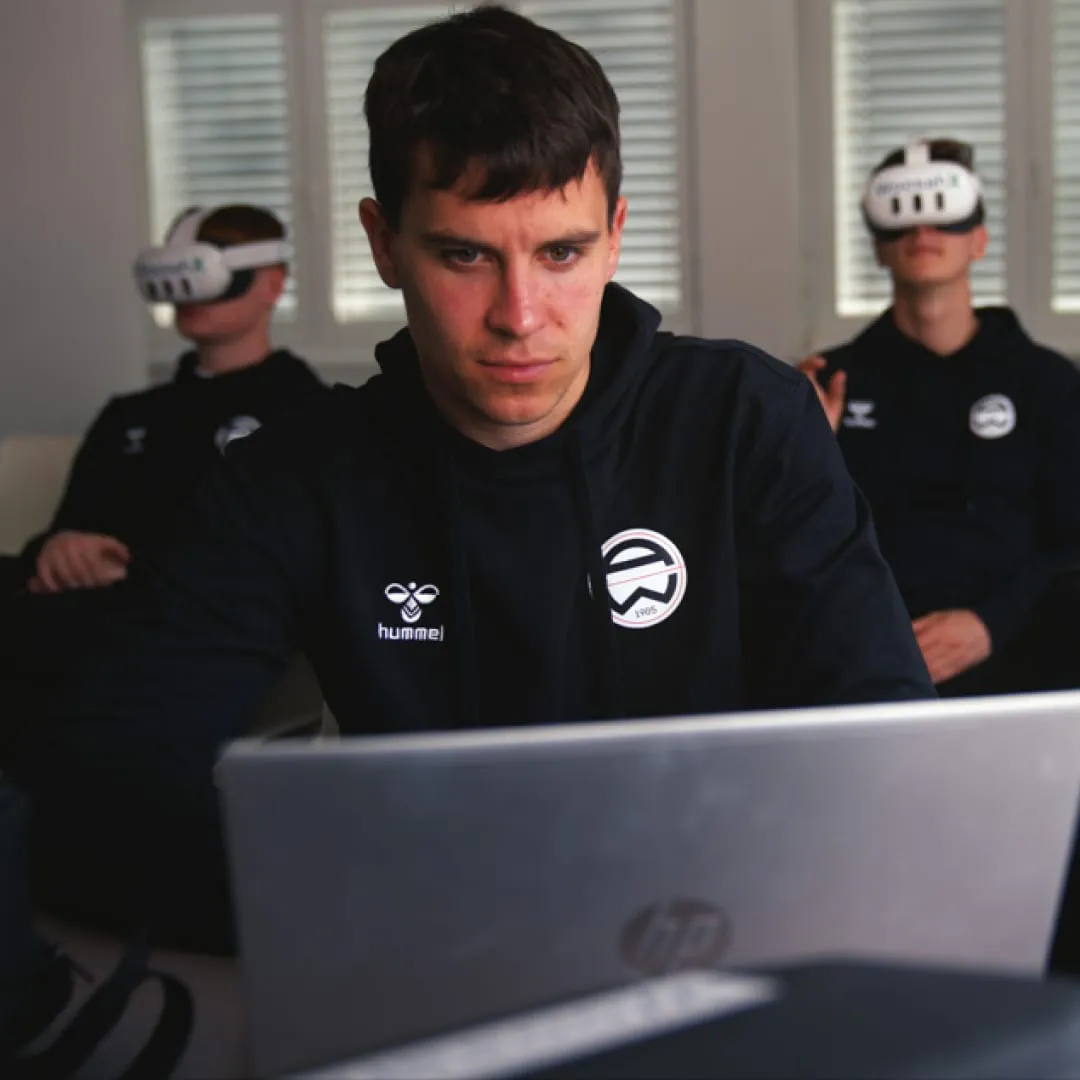 Pracovník klubu u notebooku a sportovci trénující s brýlemi pro virtuální realitu